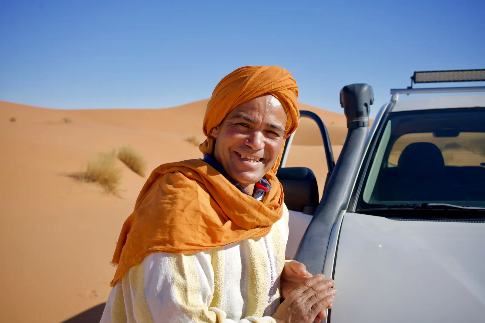 Homme souriant au chèche orange à côté d’un 4x4 blanc dans les dunes