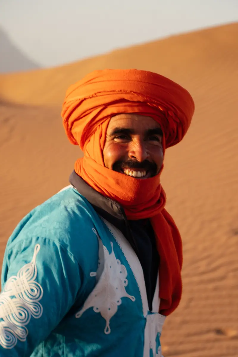 Homme souriant portant un chèche orange et un vêtement bleu devant une dune
